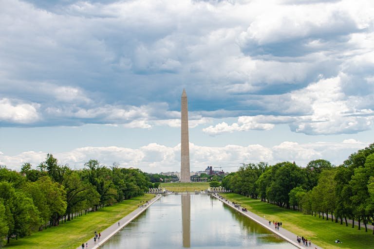 A scenic view of the Washington Monument in Washington, D.C., with reflections in the reflecting pool and a cloudy sky overhead.