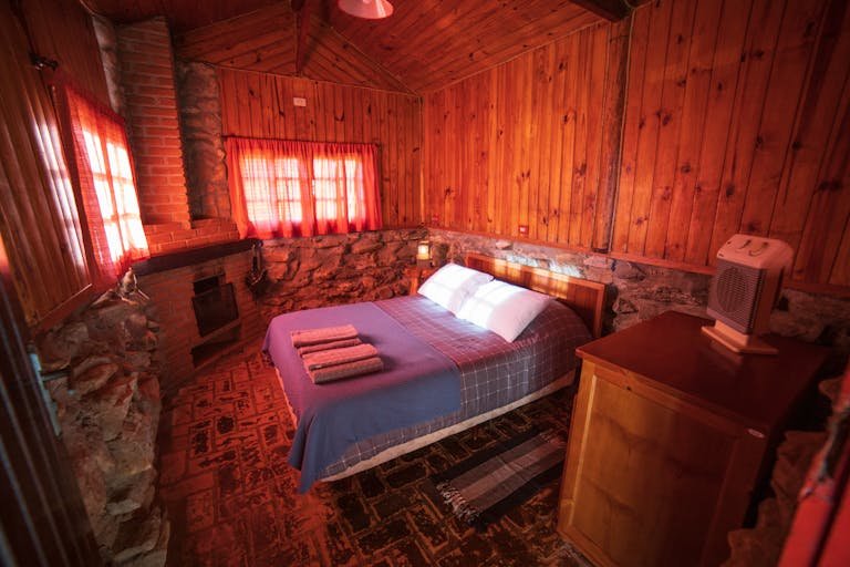 Comfortable bed with pillows placed near wooden wall decorated with stones in room with furnace of rustic house in countryside