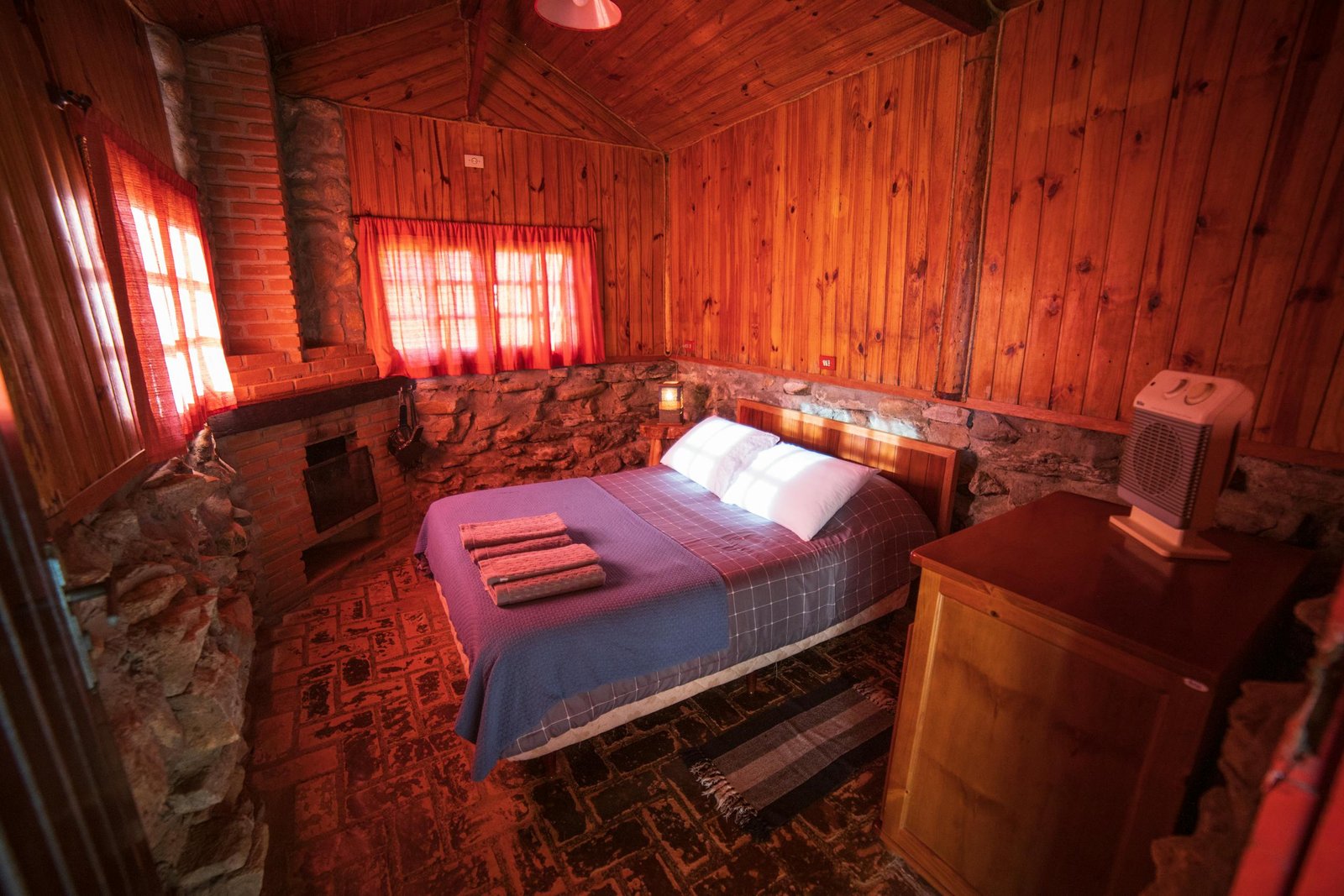 Comfortable bed with pillows placed near wooden wall decorated with stones in room with furnace of rustic house in countryside