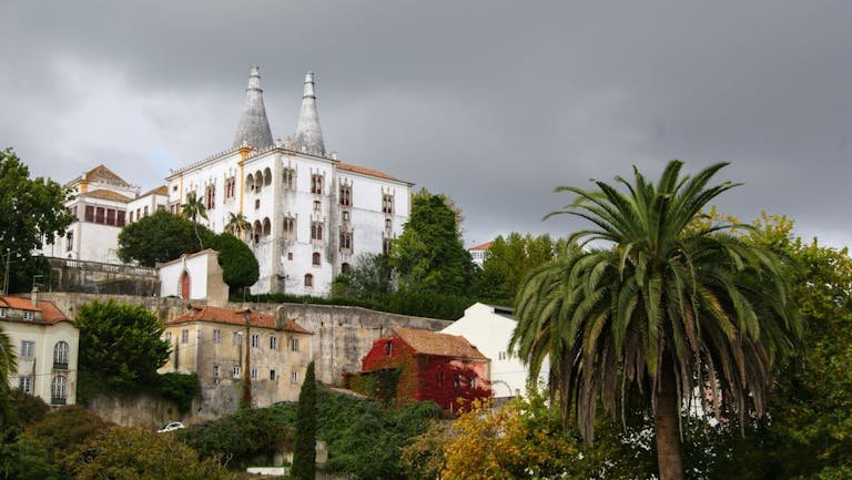 Scenic view of the National Palace of Sintra with its iconic chimneys amidst lush greenery and overcast skies.
