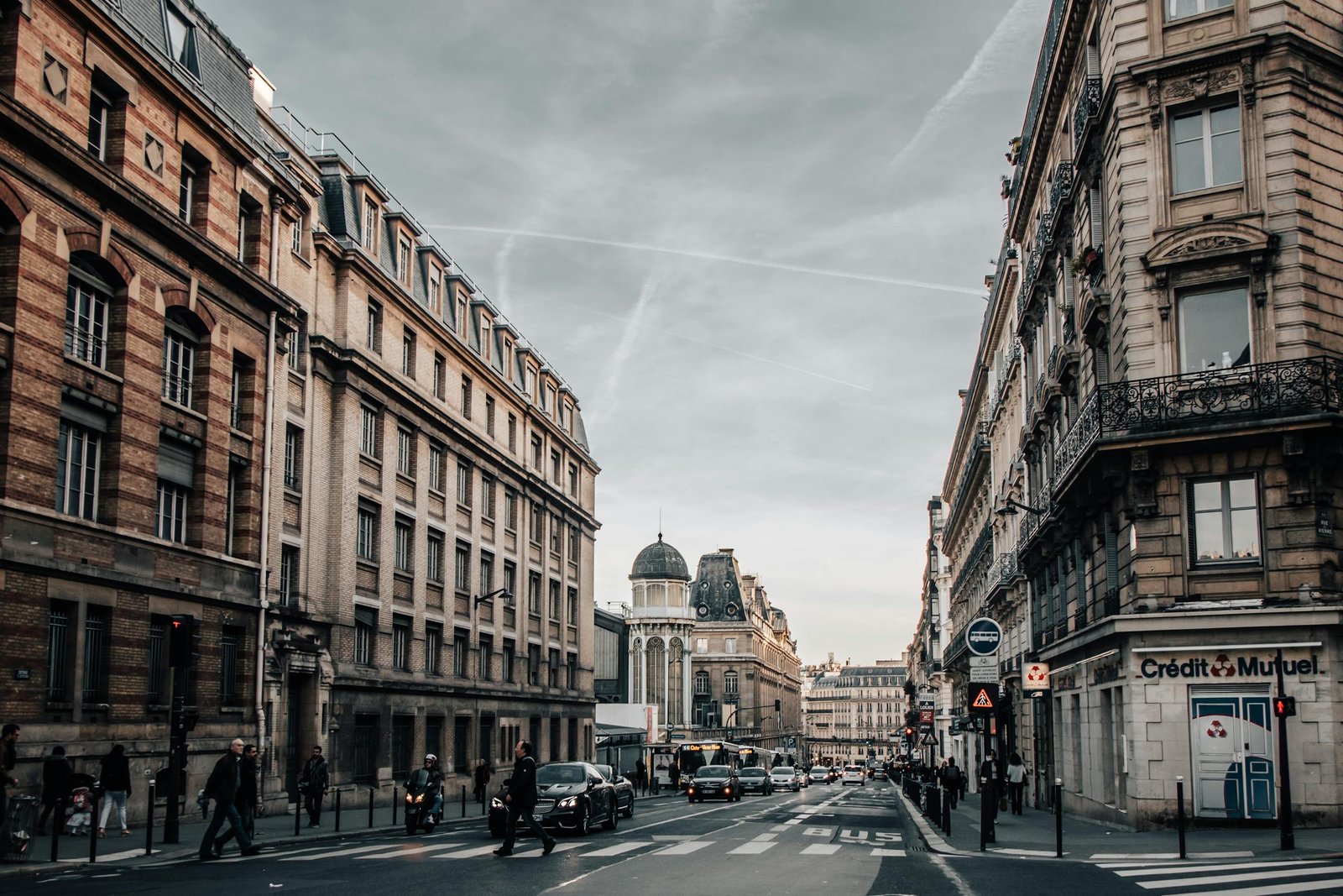 Urban street scene in Paris with classic architecture, cars, and pedestrians on an overcast day.